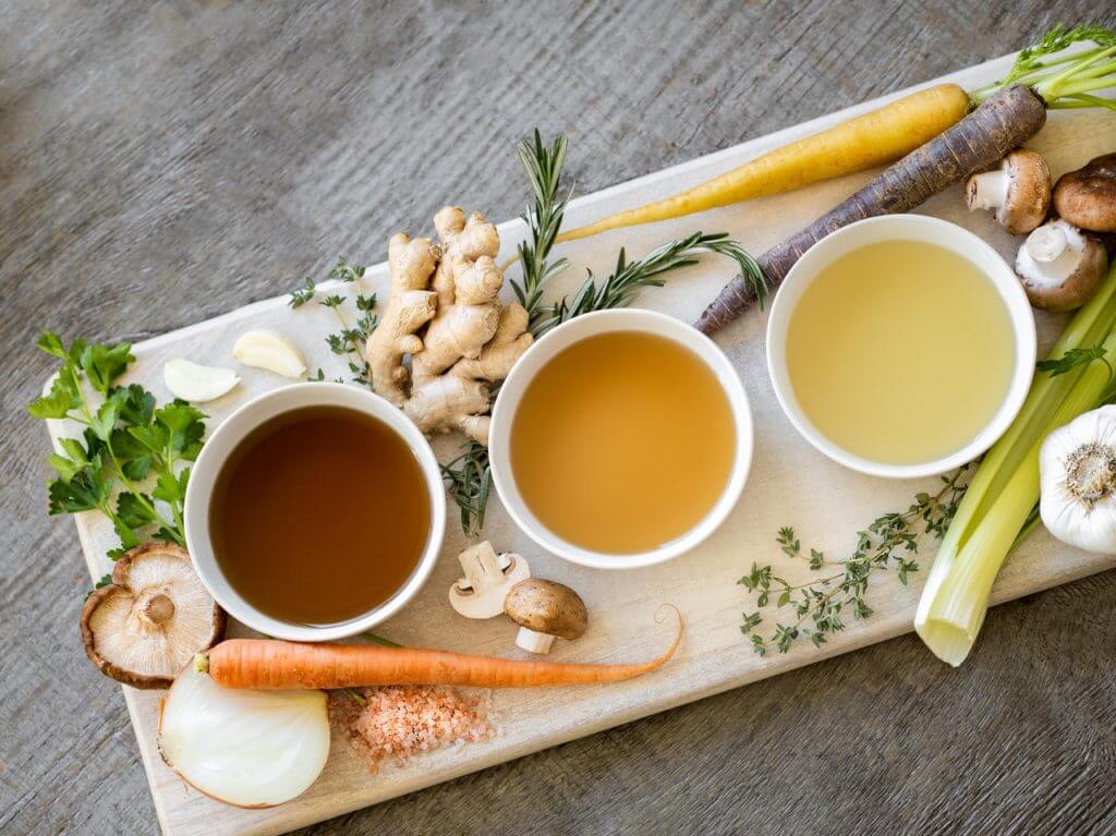 A colorful display of three cups of broth surrounded by fresh carrots and celery on a wooden table, representing healthy eating during intermittent fasting.